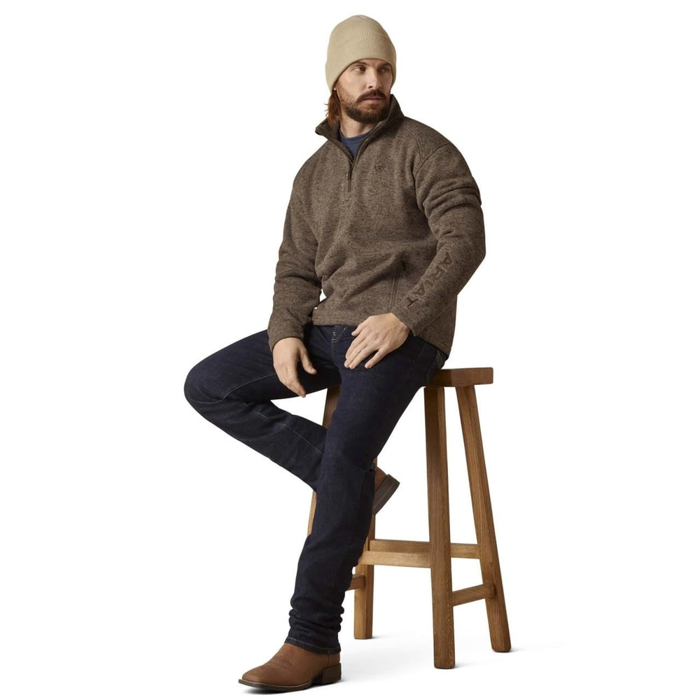 
                  
                    Man sitting on a wooden stool wearing a brown sweater, dark jeans, and a beige beanie.
                  
                