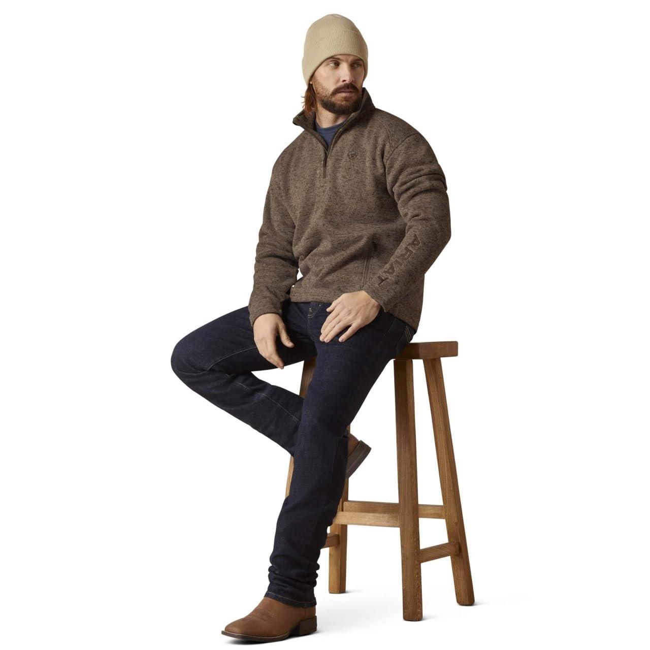 Man sitting on a wooden stool wearing a brown sweater, dark jeans, and a beige beanie.