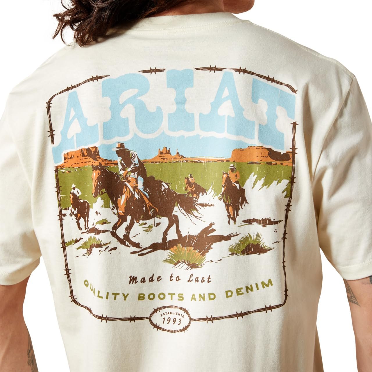 Beige t-shirt with ARIAT branding and graphic of cowboys on horses in a desert landscape.