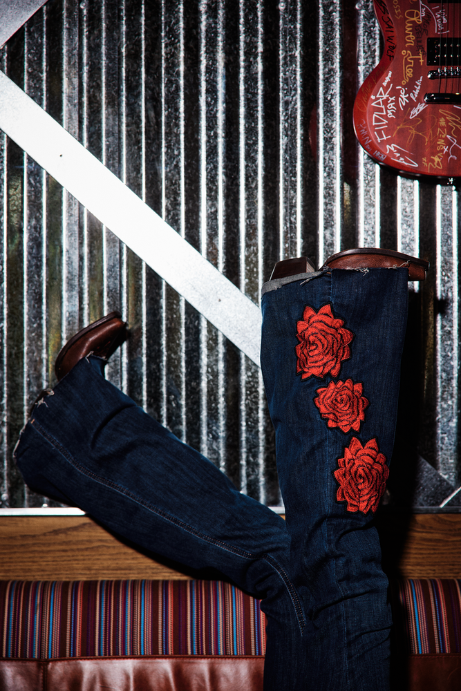 Black cowboy boots with red rose embroidery on a wooden surface with a corrugated metal wall in the background.