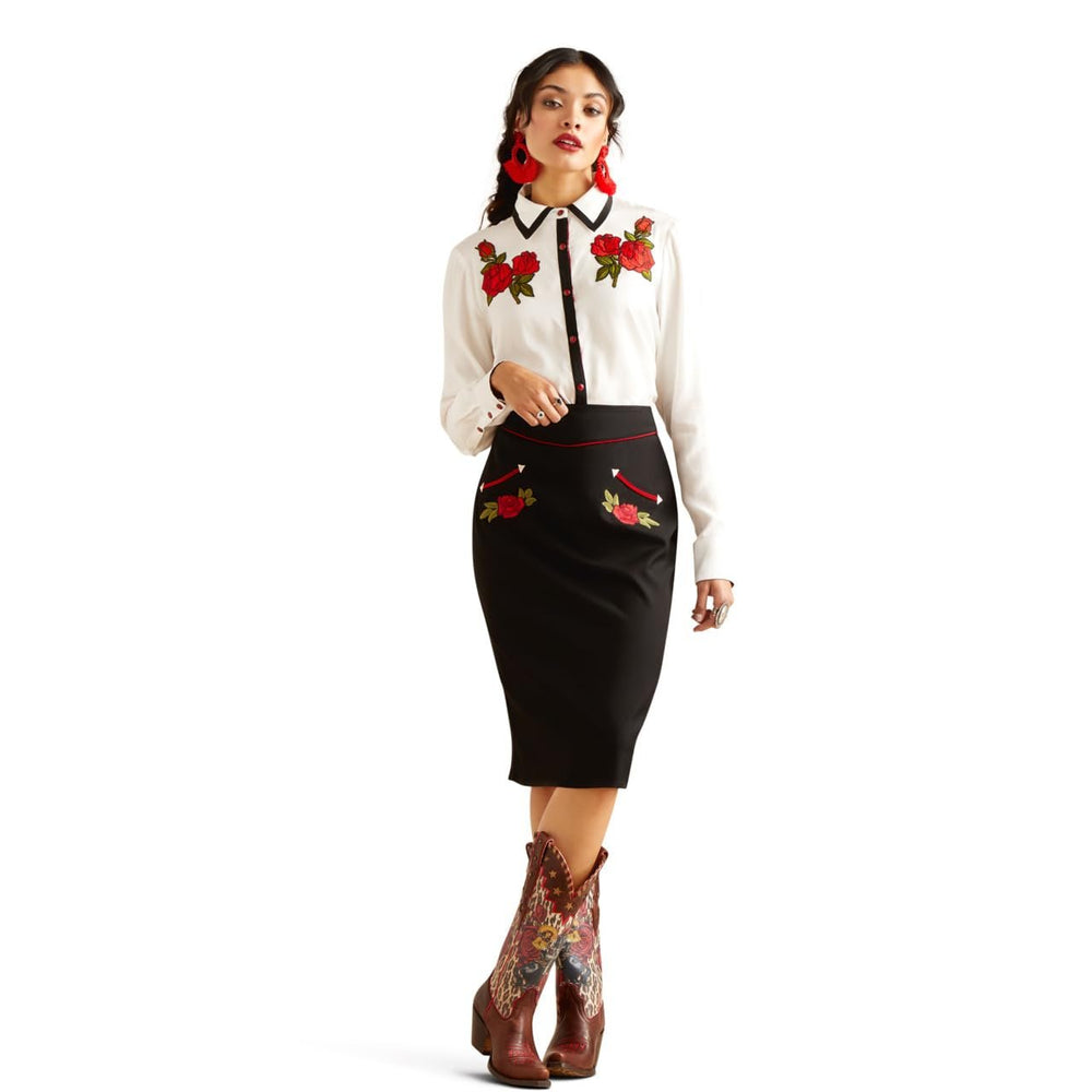 Woman wearing a white blouse with floral embroidery and a black skirt with red floral patterns, standing against a white background.