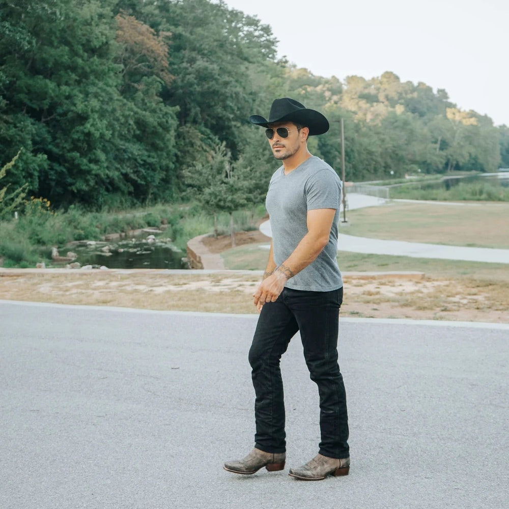 
                  
                    Man wearing the cowboy boots outdoors, paired with black jeans, a gray T-shirt, and a black cowboy hat, showcasing a modern Western look in a natural setting.
                  
                