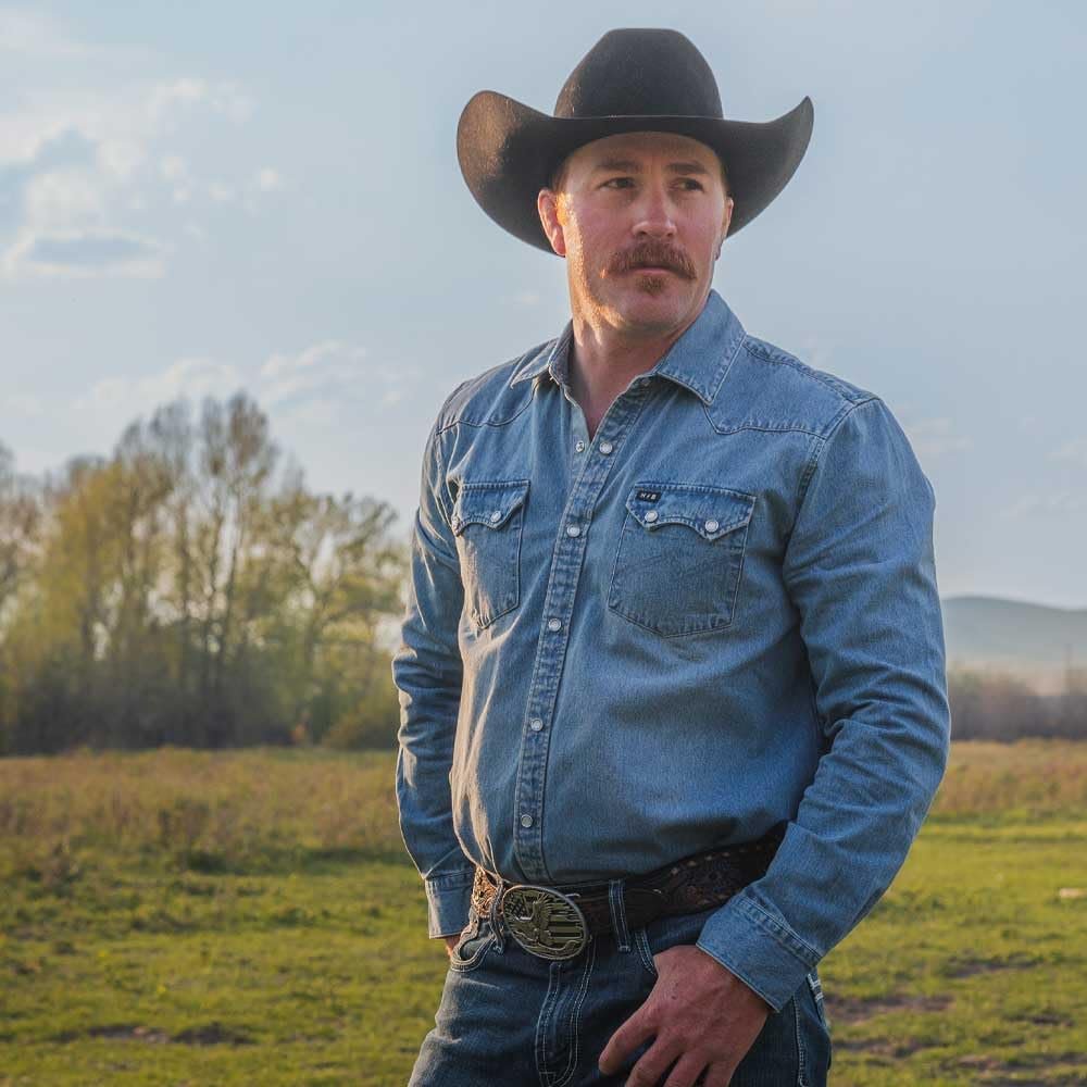 
                  
                    Man wearing a cowboy hat and blue denim shirt standing in an open field.
                  
                
