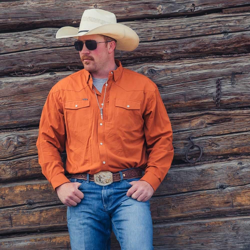 
                  
                    Man wearing an orange shirt, blue jeans, and a cowboy hat against a wooden wall.
                  
                