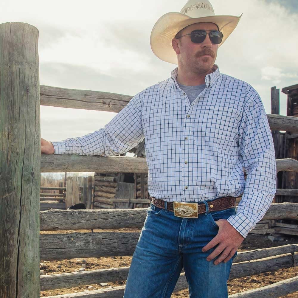 Man in cowboy hat and sunglasses standing next to a wooden fence on a ranch.