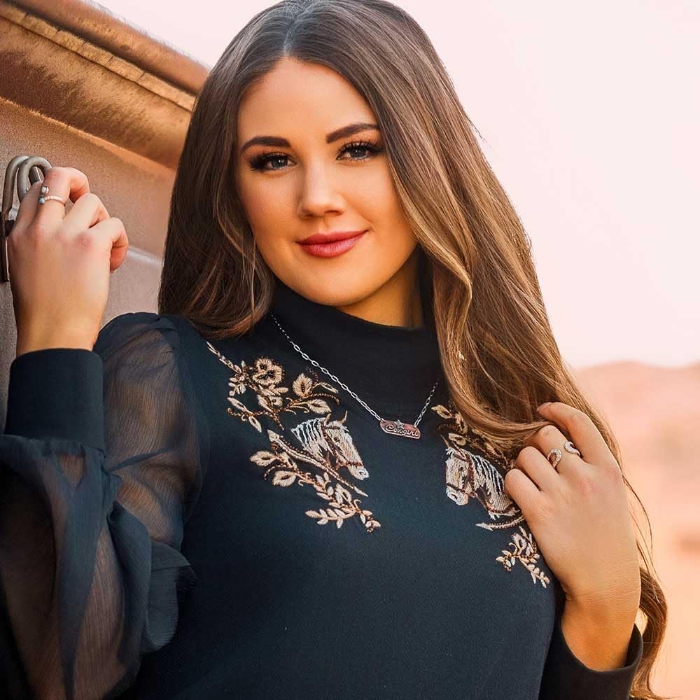 
                  
                    Woman wearing a black top with floral embroidery, standing outdoors.
                  
                