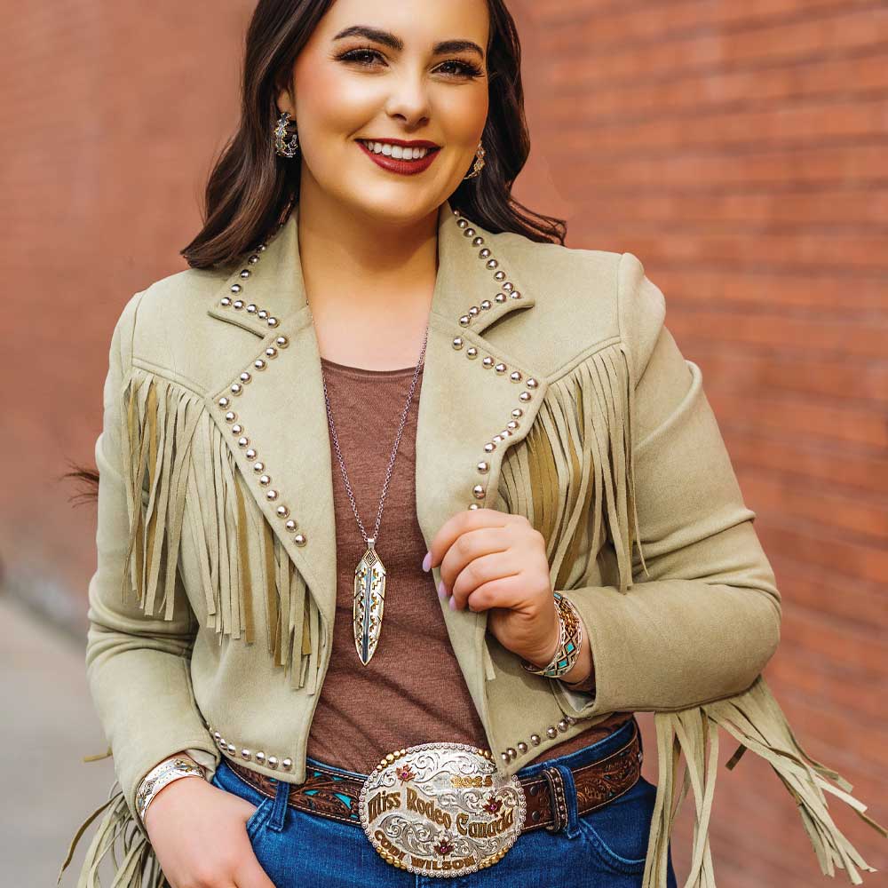 
                  
                    Woman wearing a beige fringe jacket with decorative elements against a brick wall.
                  
                