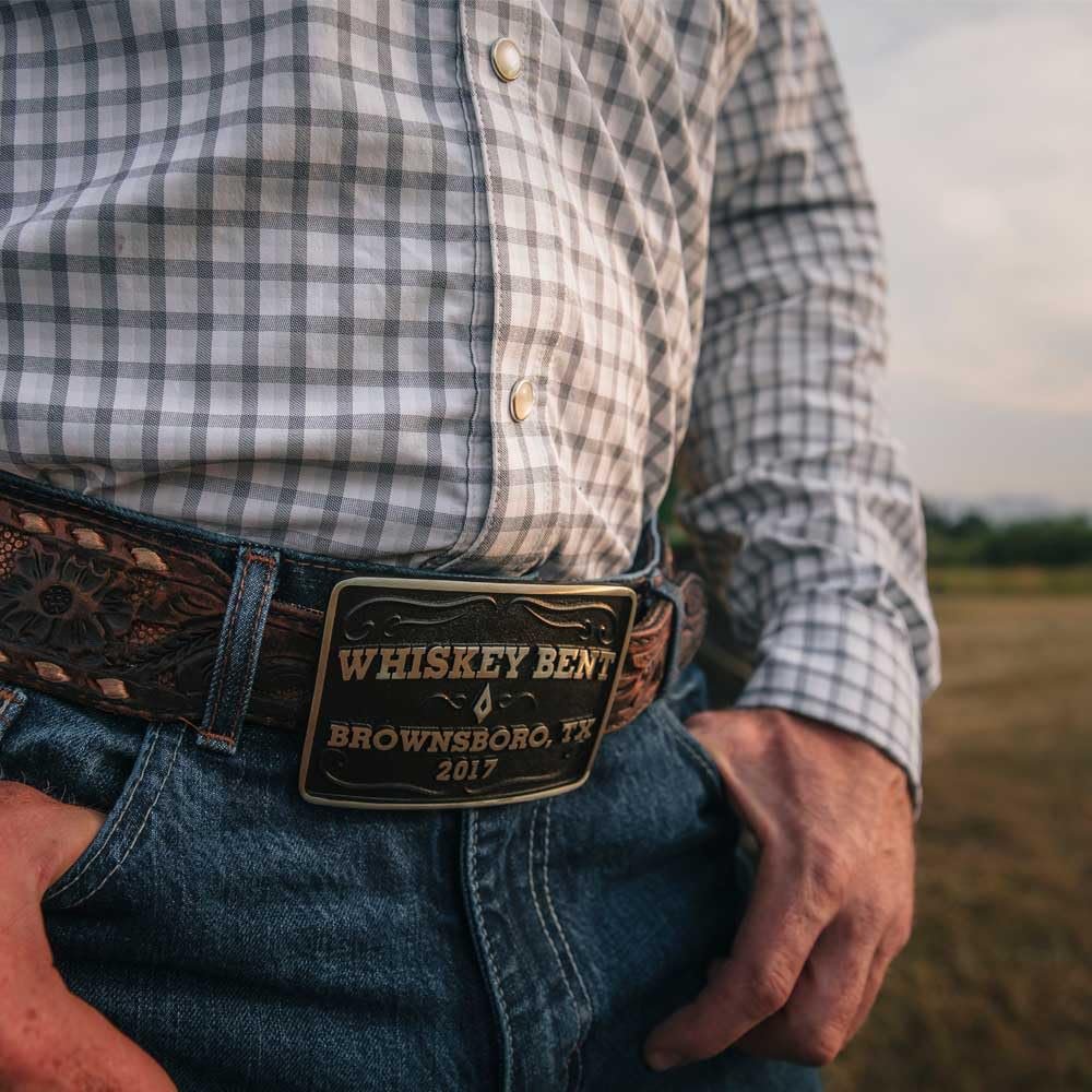 
                  
                    Man wearing Brownsboro Whiskey Bent Western Belt Buckle – A cowboy in blue jeans and a plaid shirt showcasing the Whiskey Bent western belt buckle on a tooled leather belt.

                  
                