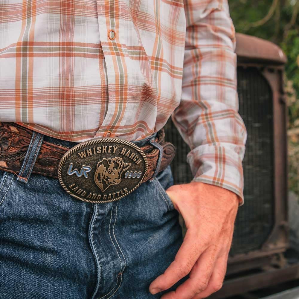 
                  
                    Man wearing Whiskey Ranch Western Belt Buckle – A close-up of a cowboy wearing a plaid shirt and jeans, showcasing the Whiskey Ranch men's western belt buckle on a tooled leather belt.
                  
                
